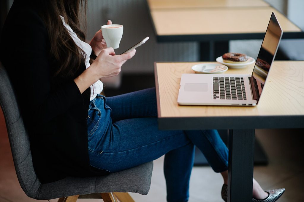 Woman at a cafe table using a smartphone with a laptop beside her