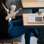 Woman at a cafe table using a smartphone with a laptop beside her